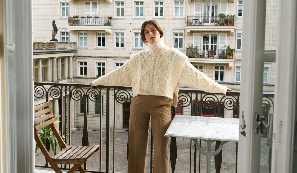 Woman in cream cable knit turtleneck sweater and brown pants on balcony with courtyard view