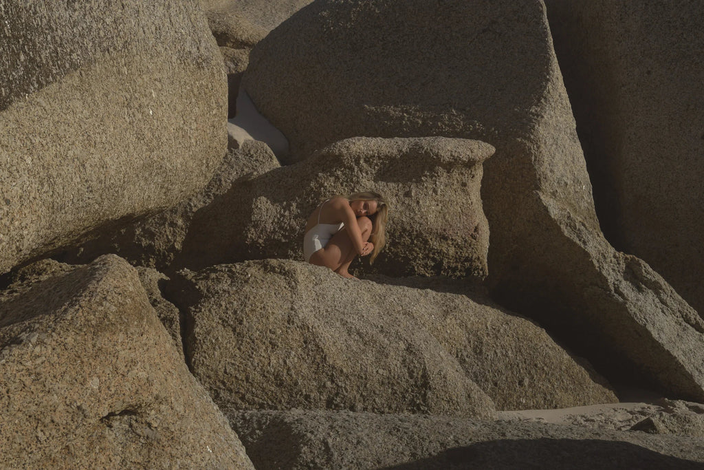 Woman in white swimsuit sitting curled up on large textured beach rocks in natural sunlight