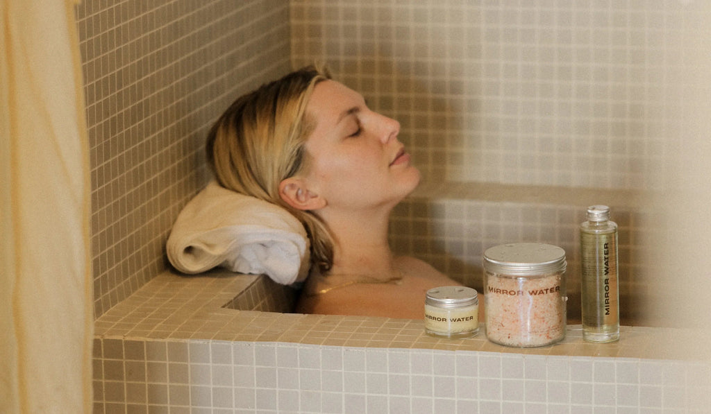 Woman relaxing in tiled bath with Mirror Water skincare products on edge