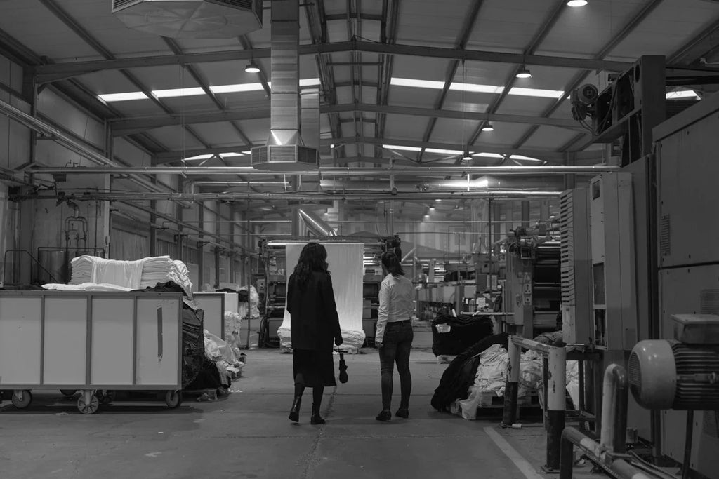 Two women walking inside a spacious textile factory with large stacks of fabric and industrial machines