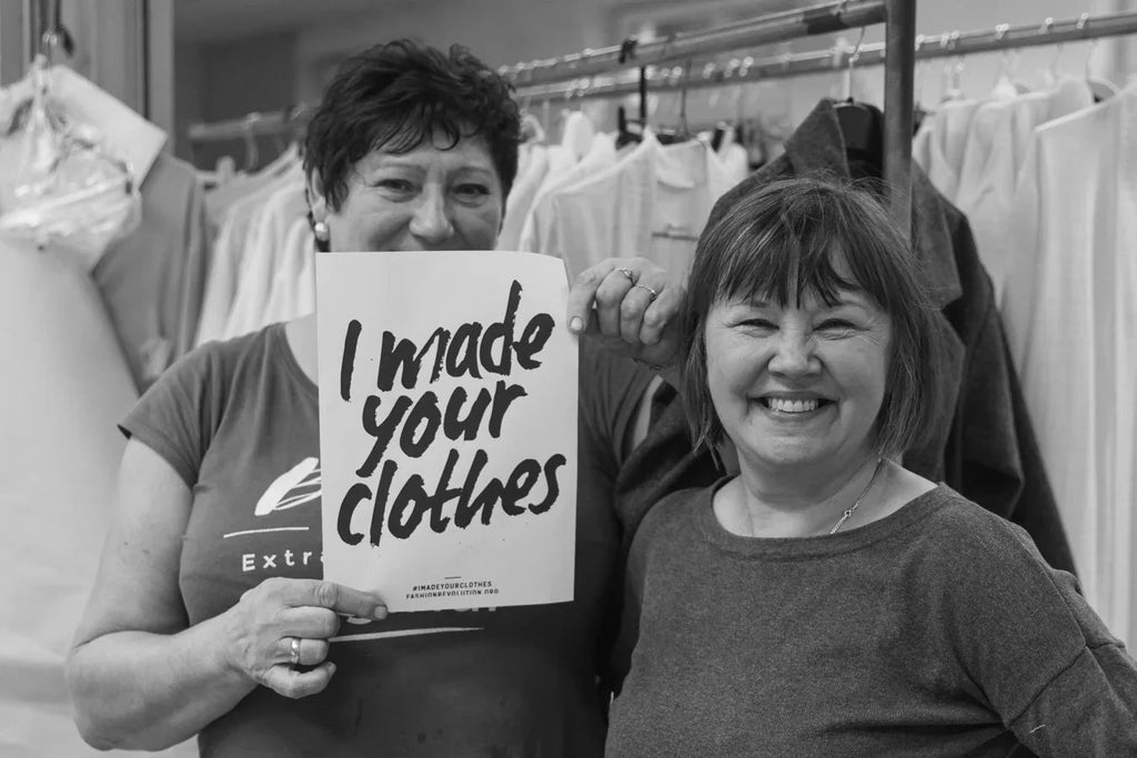Two women in a clothing workshop, one holding a sign reading I made your clothes, surrounded by garments