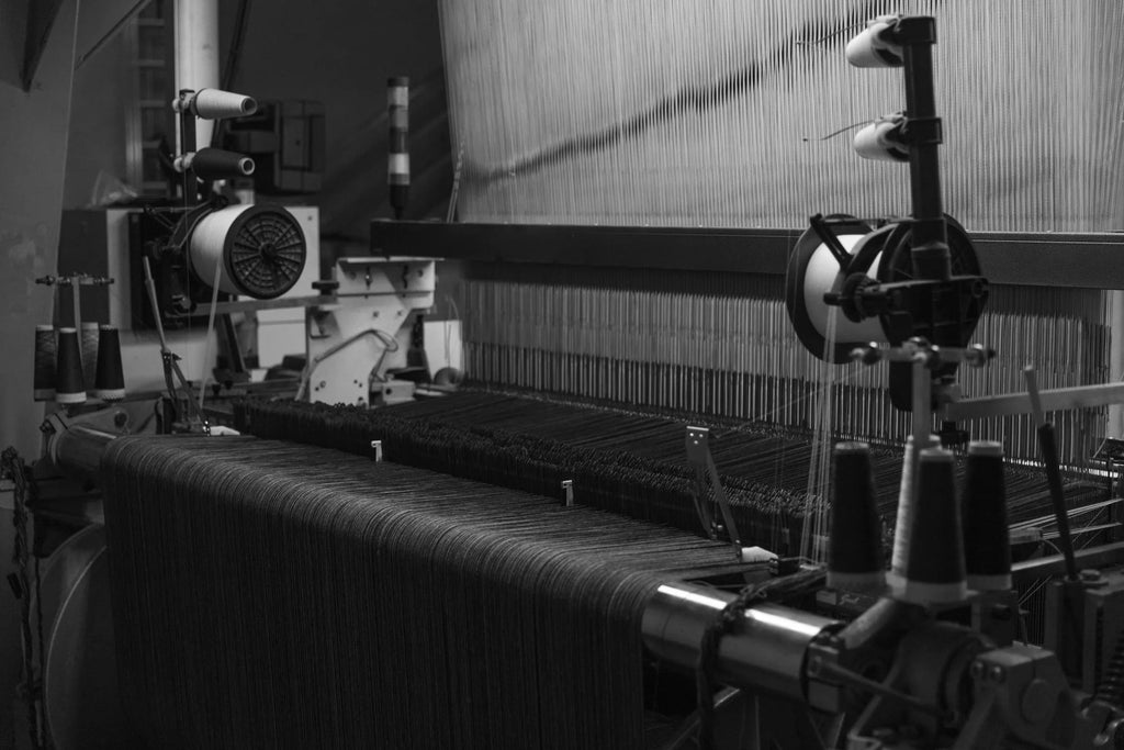 Black and white photo of industrial weaving loom with threads and yarn cones in factory setting