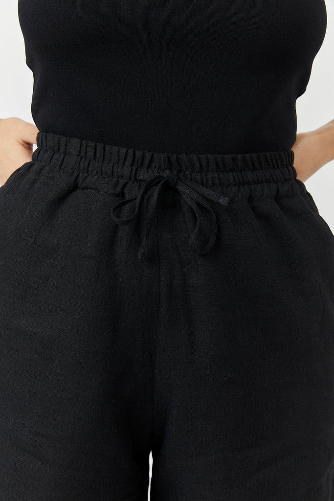 Close-up of a person wearing black linen drawstring shorts and a black top with hands on hips