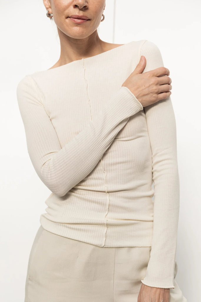 Woman wearing a cream ribbed long-sleeve top with seam detail and light beige pants on white background