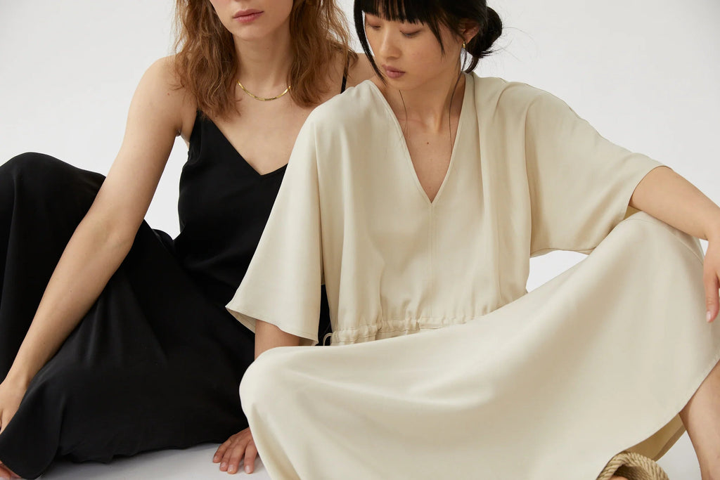 Two women sitting indoors wearing minimalist black slip dress and cream V-neck dress