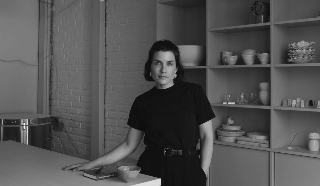 Woman in black outfit standing by table in modern room with shelves holding ceramics and glassware