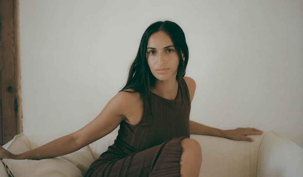 Woman with long dark hair in sleeveless brown dress sitting on white couch against plain wall